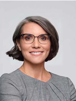 A headshot of Carmen Rojas. She has short brown hair and glasses and is wearing a gray top